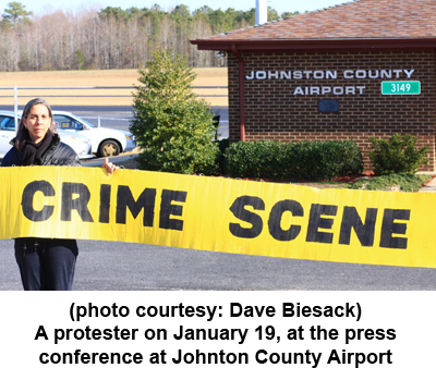 Photo of protester holding a sign reading "Crime Scene" in front of Johnston County Airport